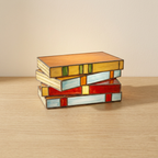 Candeeiro de livros empilhados com efeito vitral numa mesa de carvalho claro, junto a uma parede creme