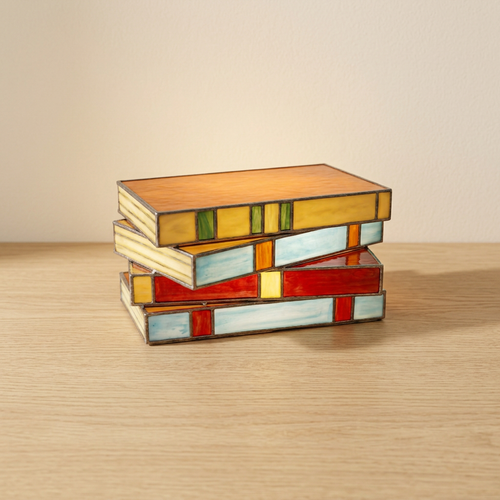 Candeeiro de livros empilhados com efeito vitral numa mesa de carvalho claro, junto a uma parede creme